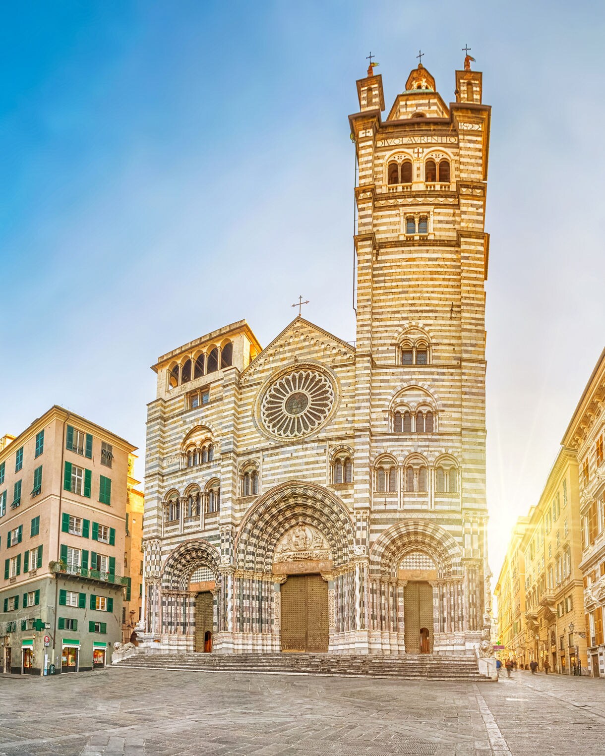 The Cathedral of San Lorenzo in Genoa, Italy, with its striped Gothic façade and twin towers illuminated by sunlight, surrounded by historic buildings.