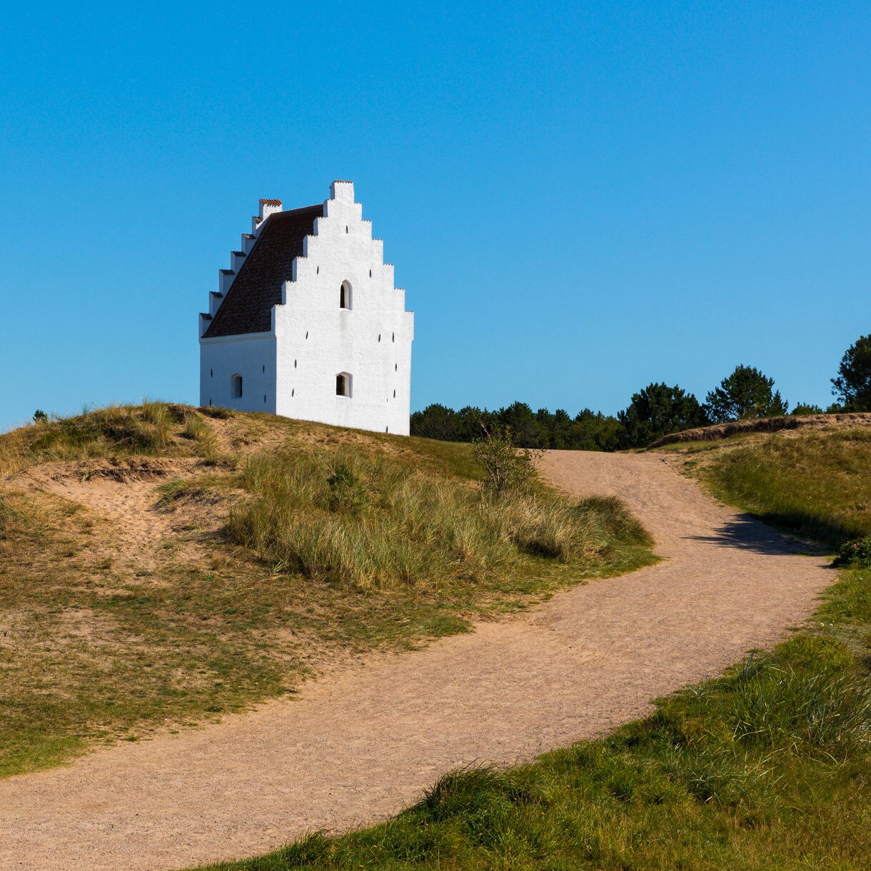 White stepped-gable Sand-Covered Church in Skagen, Denmark, standing among sandy dunes and grassy paths under a clear blue sky.