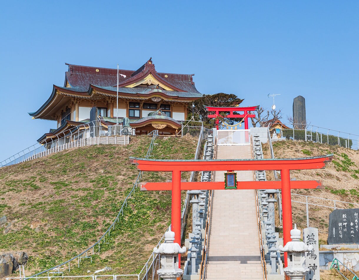 Hilltop Shinto shrine with ornate wooden architecture, reached by a long stone staircase lined with bright red torii gates and lanterns, set under a clear blue sky.