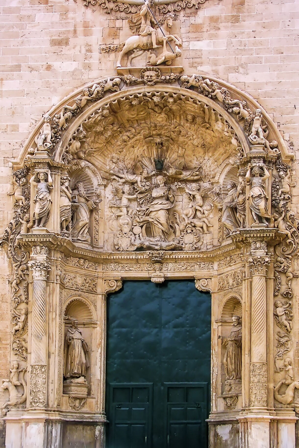 Close-up of the ornate Baroque entrance of the Church of Sant Francesc in Palma, Mallorca, featuring detailed stone carvings of saints and biblical figures surrounding a green door.