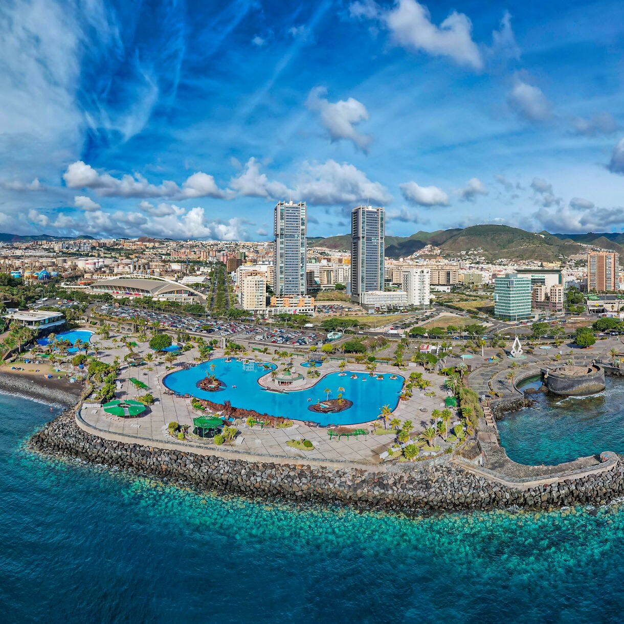 Panoramic aerial view of Santa Cruz de Tenerife, Canary Islands, showing the city’s waterfront, Auditorio de Tenerife, pools and surrounding ocean under a partly cloudy sky.
