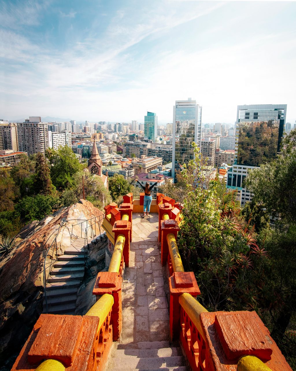 View from a bright red and yellow stairway atop Santa Lucía Hill overlooking Santiago’s modern skyline, with trees and rocky paths surrounding the lookout.