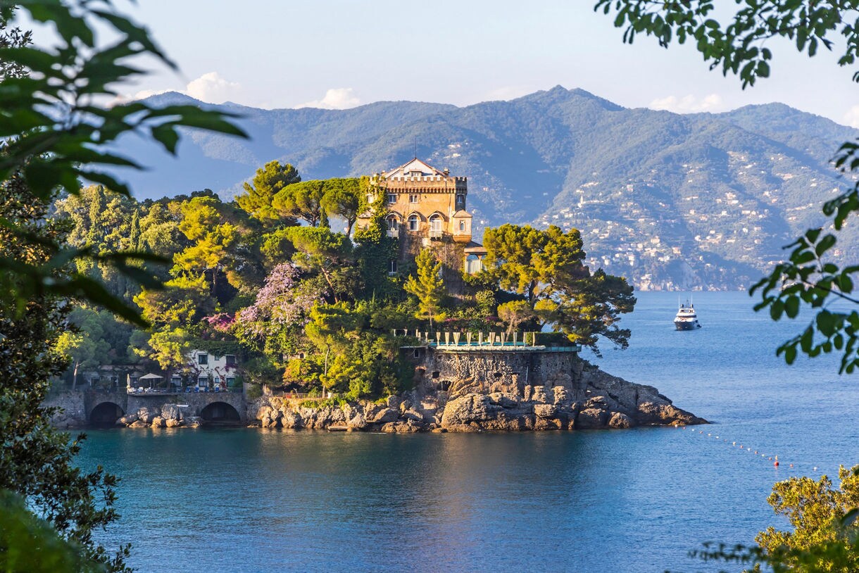 Colorful seaside town with palm trees and layered hillside villas overlooking a calm pebbled beach.