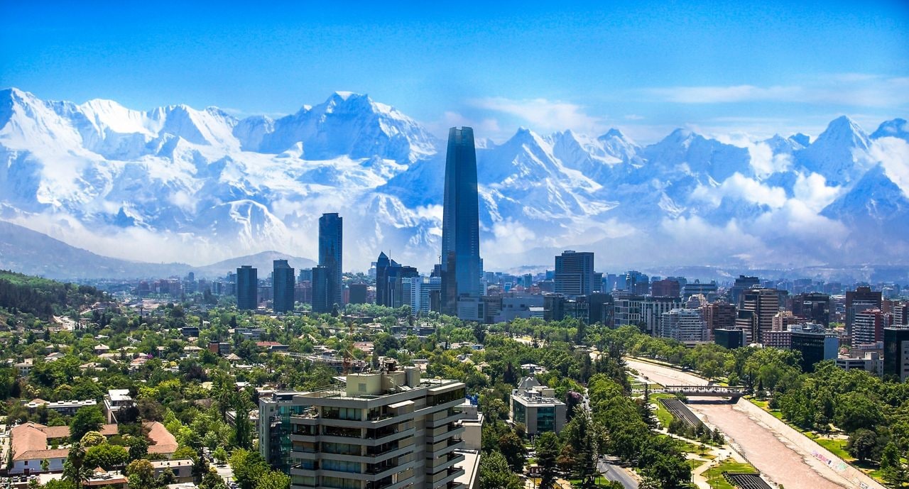A panoramic view of Santiago with modern skyscrapers and leafy neighborhoods set against massive snow-covered Andes mountains.