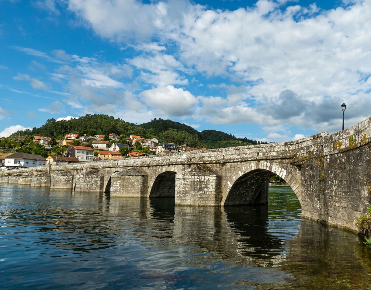 Old stone arch bridge crossing a reflective river with a hillside village of red-roofed houses and green trees in the background under a partly cloudy sky.