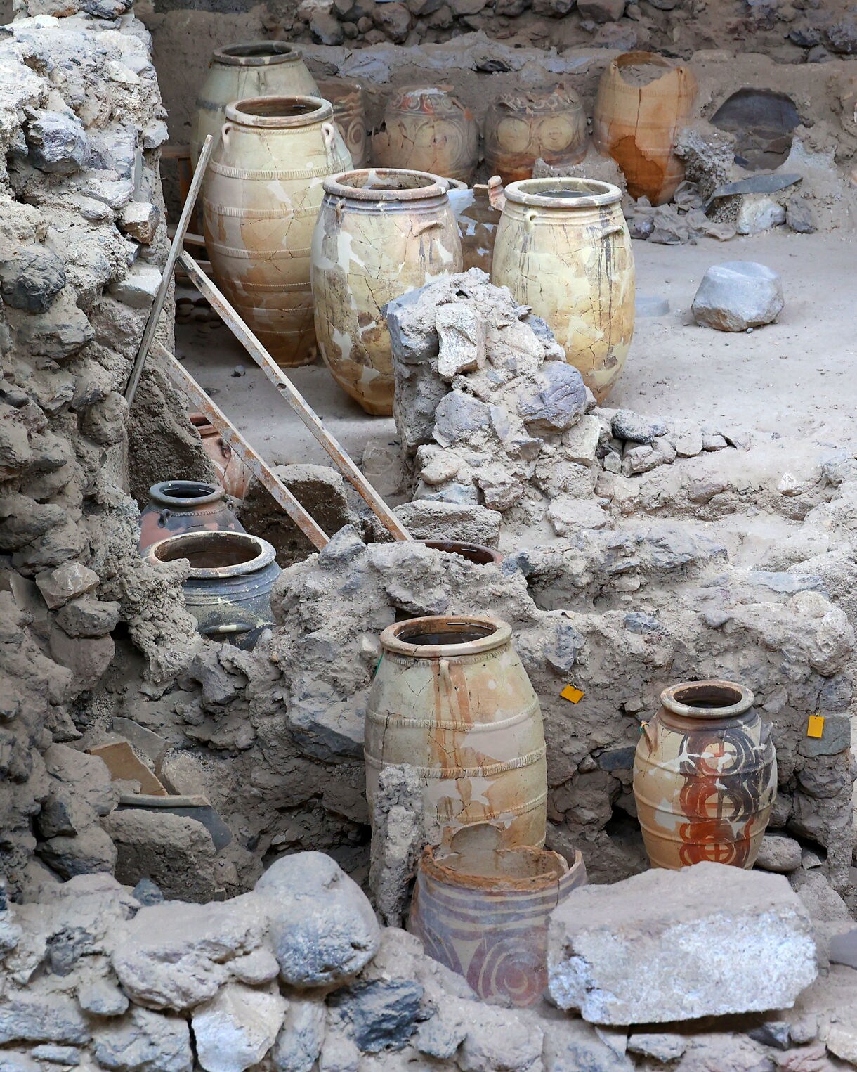 Excavated ruins of Akrotiri on Santorini with large clay storage jars scattered among stone walls and rubble.