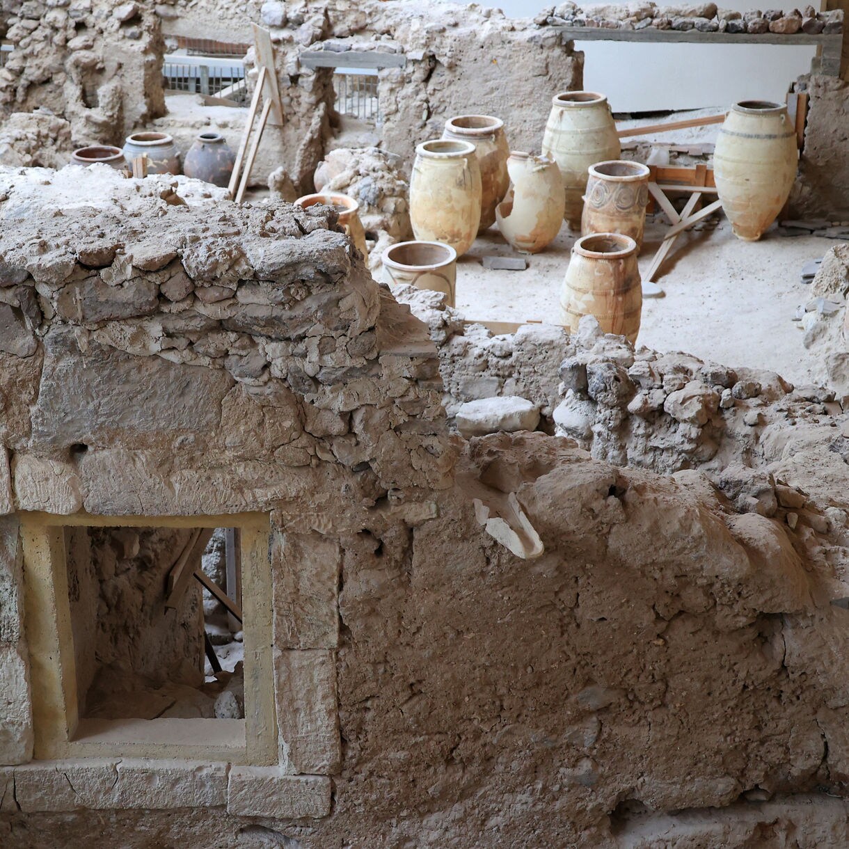 Excavated ruins of Akrotiri on Santorini showing stone walls, doorways, and clay storage jars arranged in a partially preserved room.