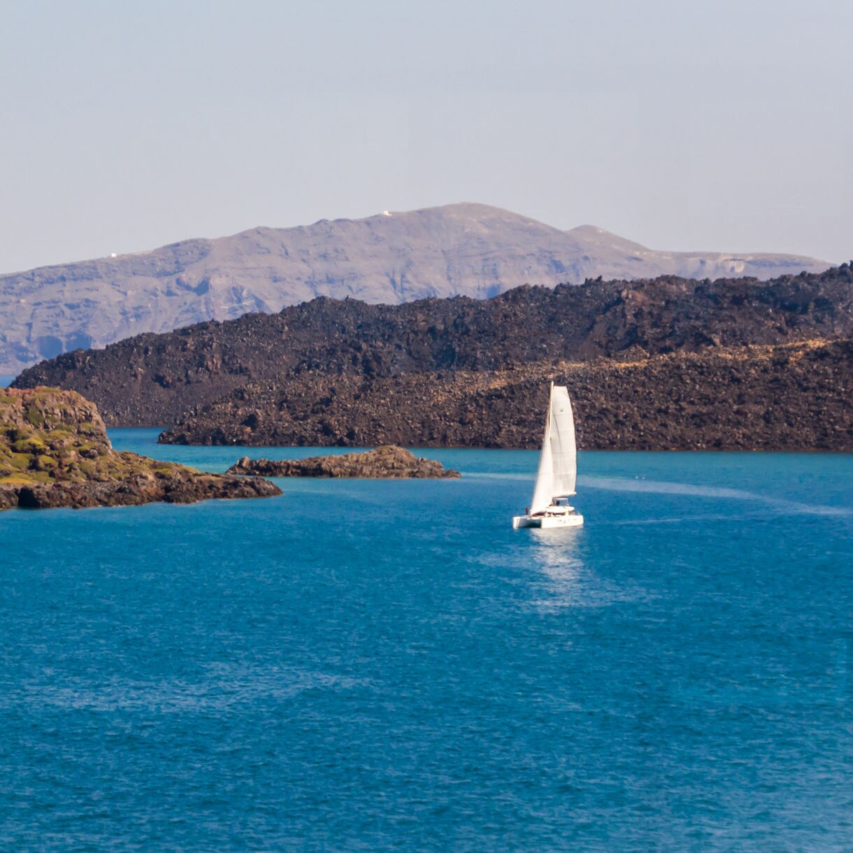 White catamaran gliding through the turquoise waters of Santorini’s caldera with rugged volcanic islands and distant cliffs in the background.