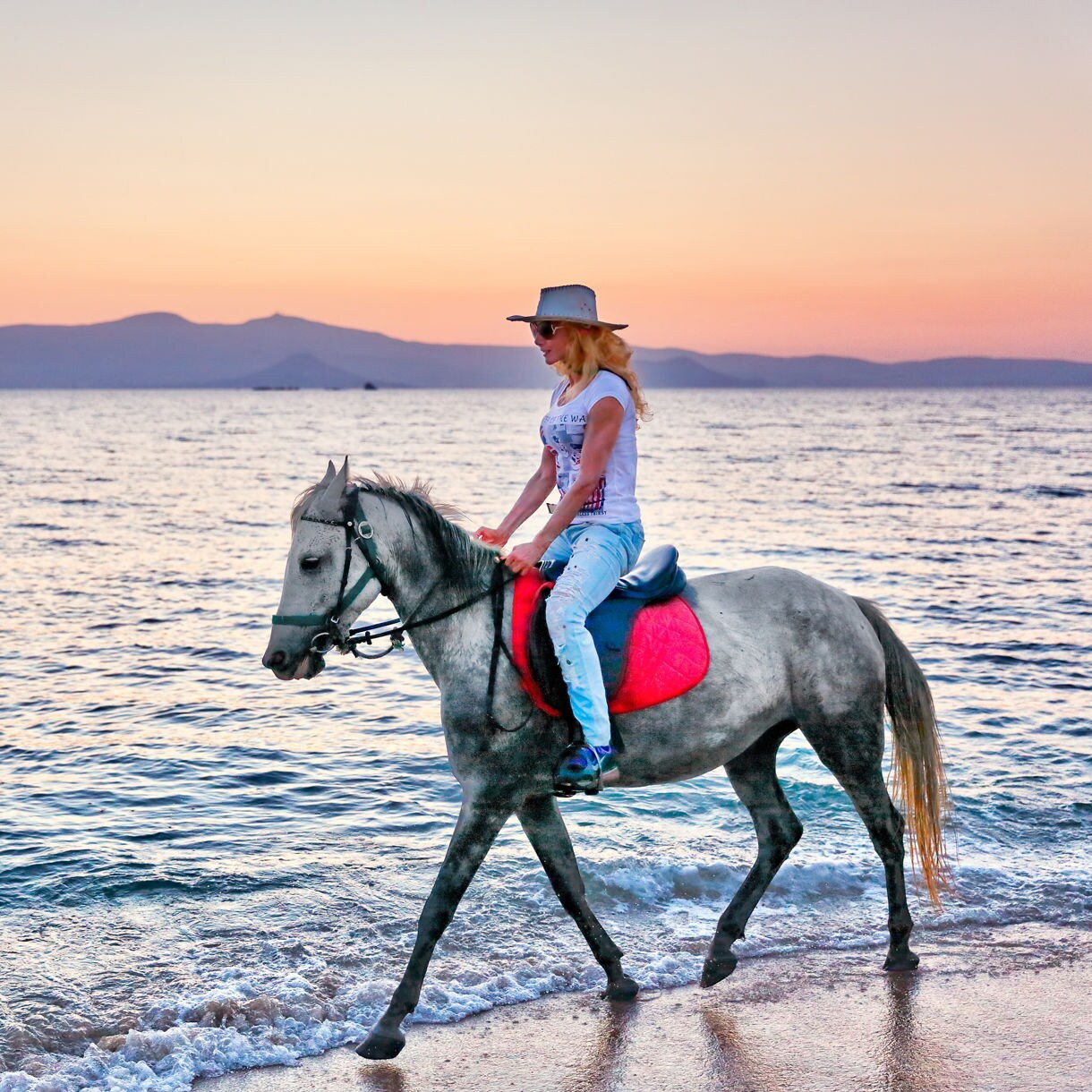 Woman in a hat riding a gray horse with a red saddle pad on the beach as the sun sets over the Aegean Sea.