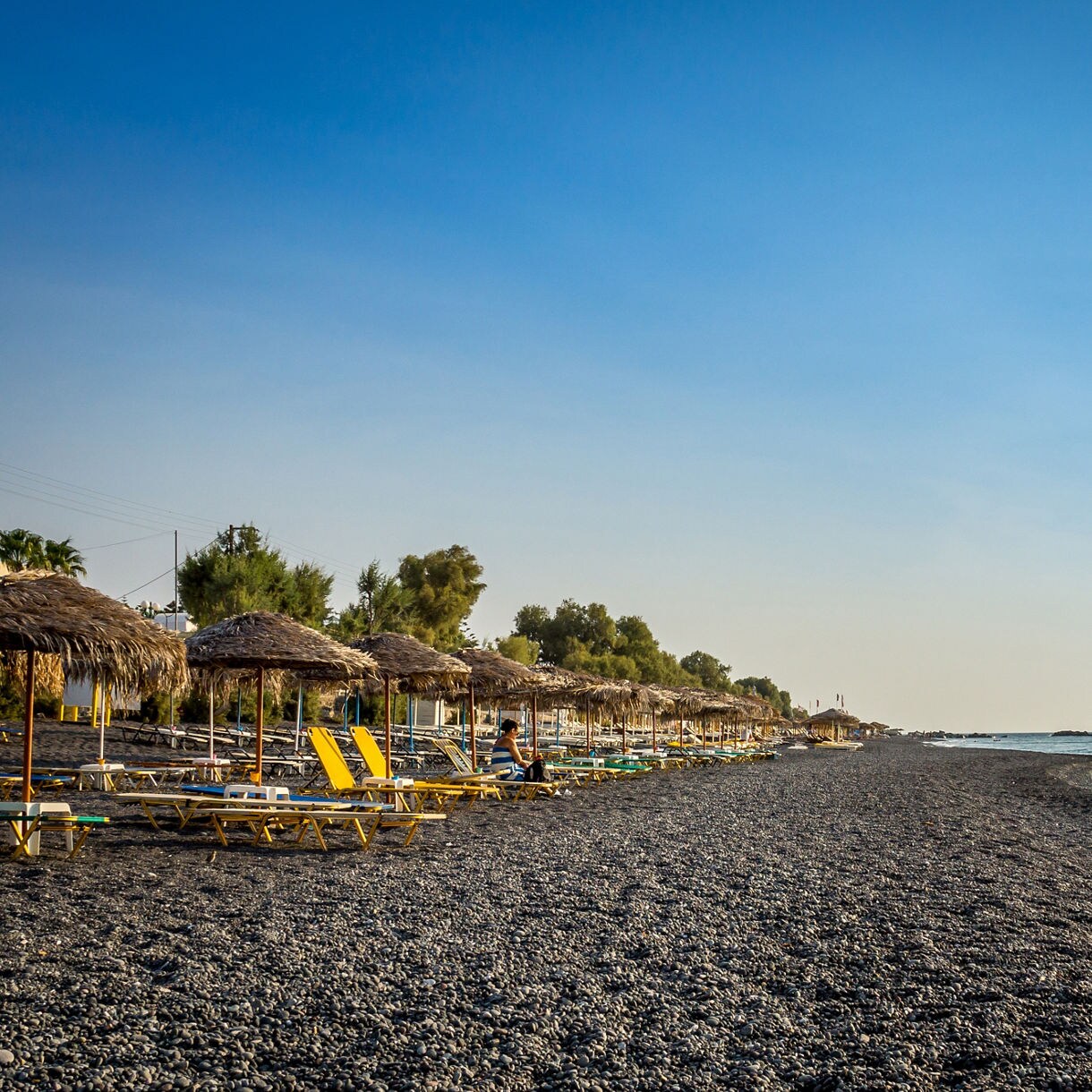 Wide view of Kamari Beach in Santorini with rows of straw umbrellas and lounge chairs on dark volcanic pebbles beside calm blue water.