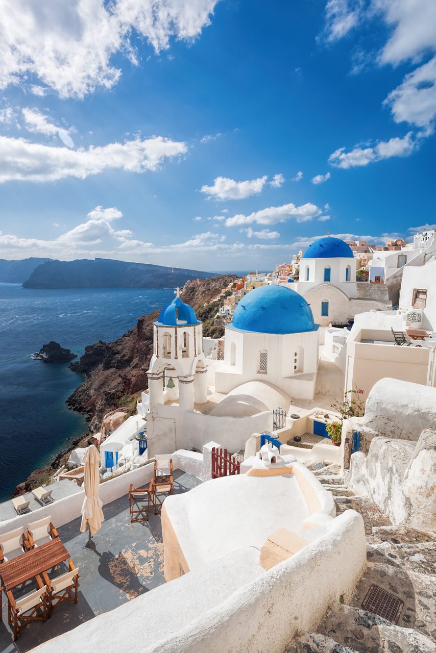 Vertical view of Santorini’s cliffside village with white buildings, blue-domed churches, and the deep blue Aegean Sea under a bright sky.