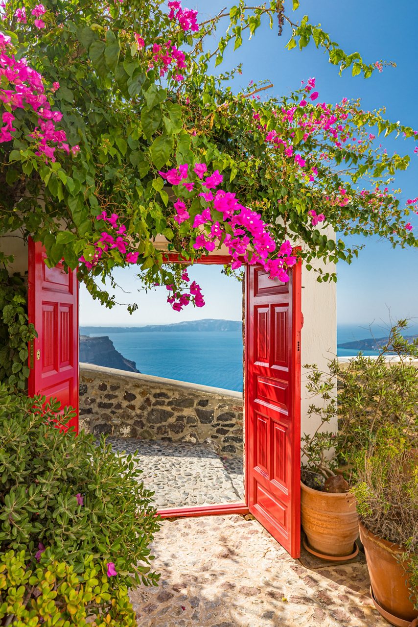 Bright red doorway framed with pink bougainvillea flowers opening to a stone patio with a view of the blue Aegean Sea and distant cliffs.