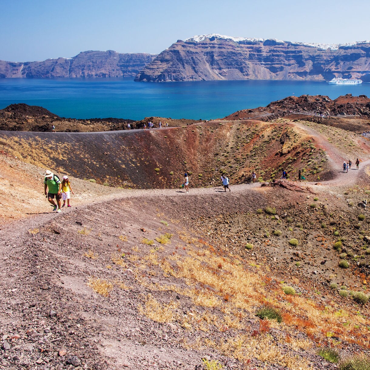 Tourists walking along winding trails on Santorini’s volcanic island, with red and black rocky terrain overlooking the blue Aegean Sea and distant cliffs.