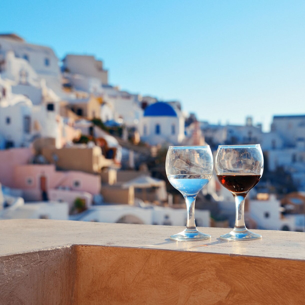 Two glasses of wine on a terrace overlooking Santorini’s whitewashed houses with blue domes and pastel facades under a clear sky.