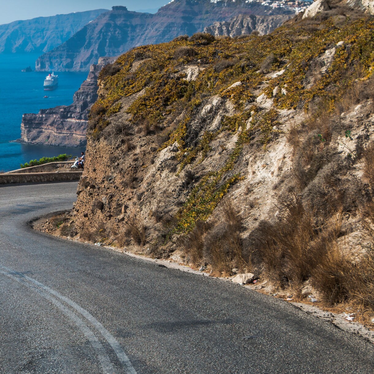 Curving coastal road in Santorini hugging a cliffside, overlooking steep volcanic slopes and the Aegean Sea with cruise ships in the distance.