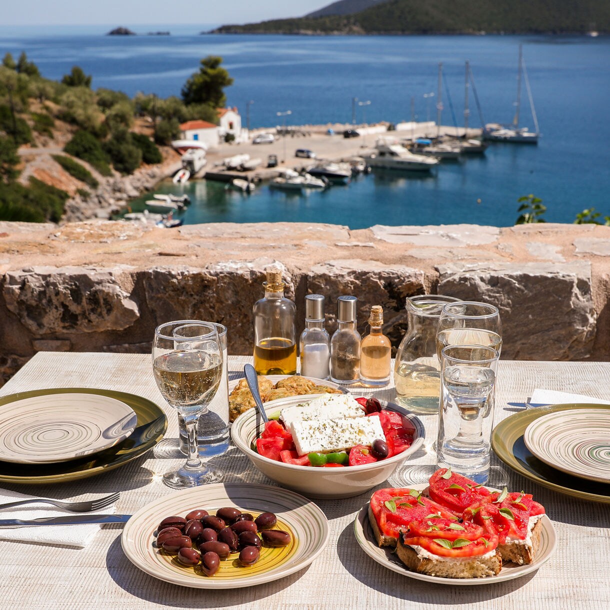 Outdoor table set with Greek dishes including olives, tomato bruschetta, and a salad with feta cheese, with a view of boats docked in a blue bay.
