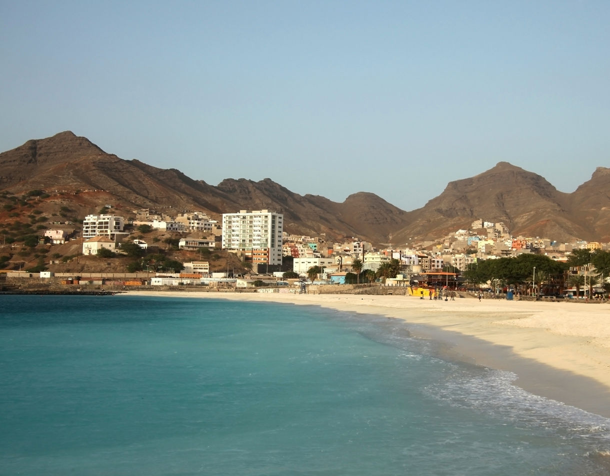 Coastal view of São Pedro Beach with pale sand, gentle turquoise waves, scattered beachgoers and a hillside town backed by steep rocky mountains under a clear sky.
