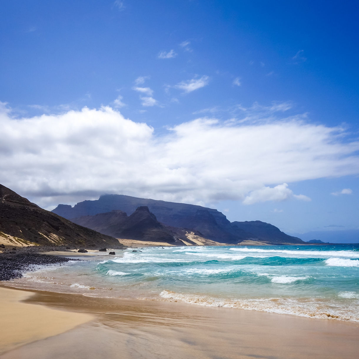 Wide coastal view of a rugged beach on São Vicente Island with soft sand, rocky slopes, turquoise surf and distant volcanic mountains under a bright sky with scattered clouds.