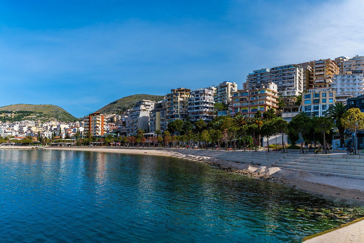 Wide panoramic view of Sarandë’s waterfront in Albania showing terraced apartment buildings climbing green hills above a long beach, calm turquoise water and a tree-lined promenade.