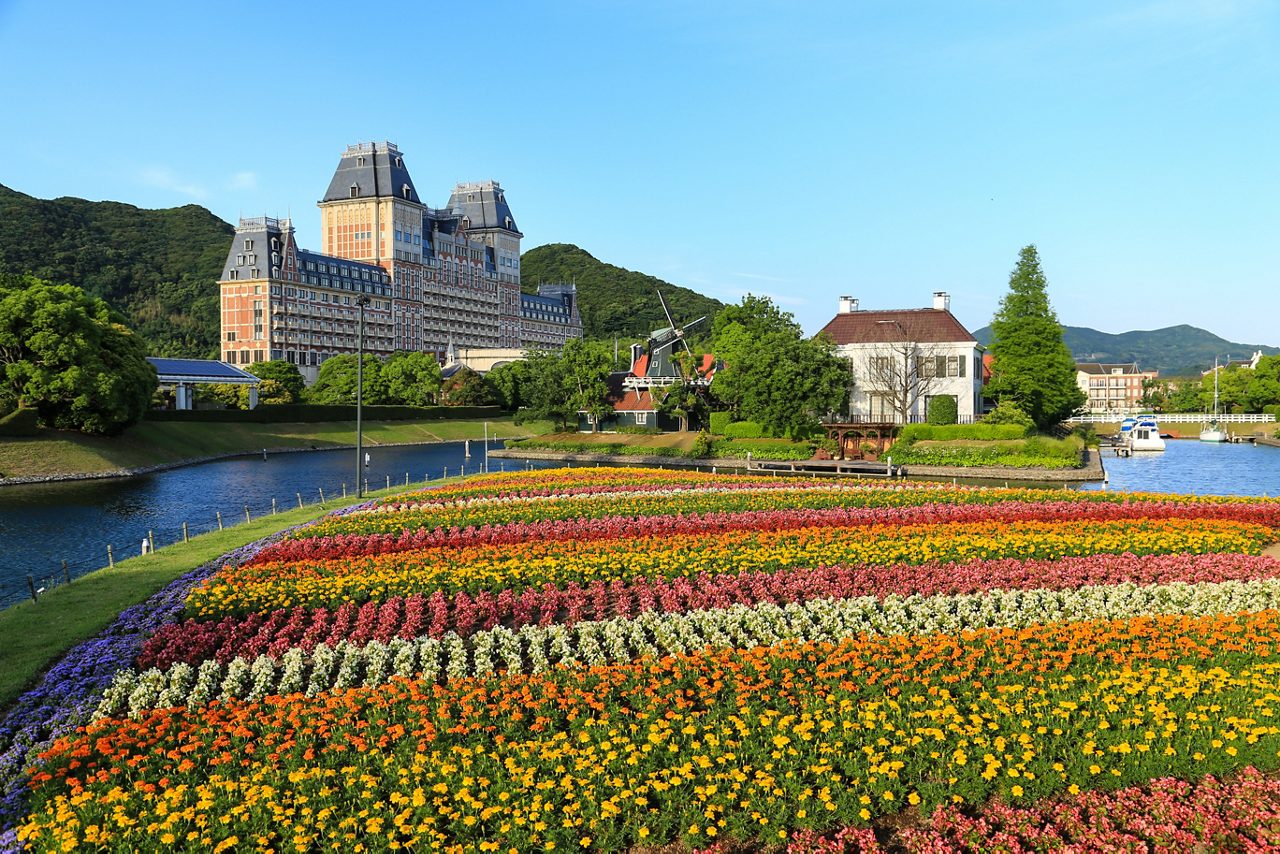 Rows of vibrant flowers lining a canal with Dutch-style buildings and a windmill in Huis Ten Bosch theme park, Sasebo, Japan.