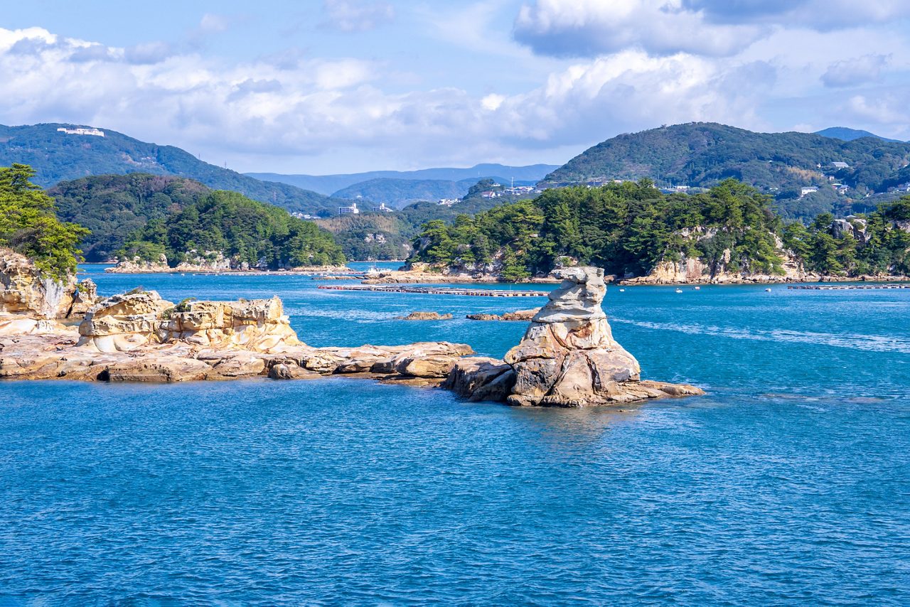 Weathered rock formations jutting from turquoise water with forested coastline and mountains behind.