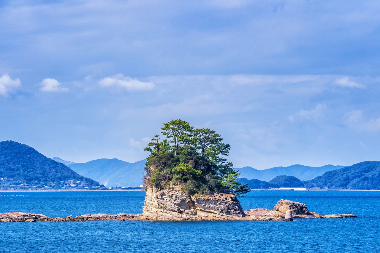 Rocky island topped with pine trees surrounded by blue water, with forested mountains in the distance.