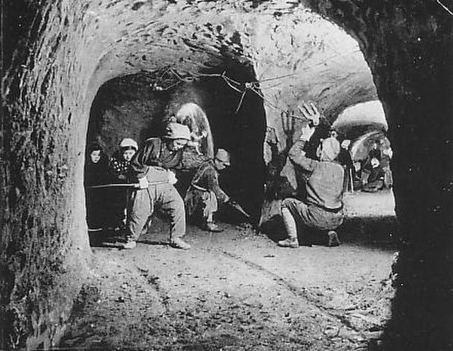 Black and white photo of wartime mannequins reenacting a scene inside an underground air raid shelter with arched tunnels.