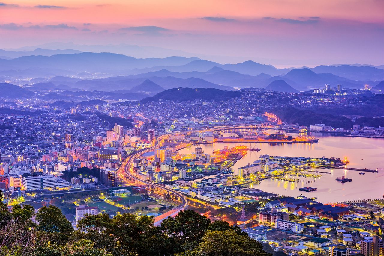 Aerial view of Sasebo at dusk with glowing city lights, winding roads and a calm harbor surrounded by hills.