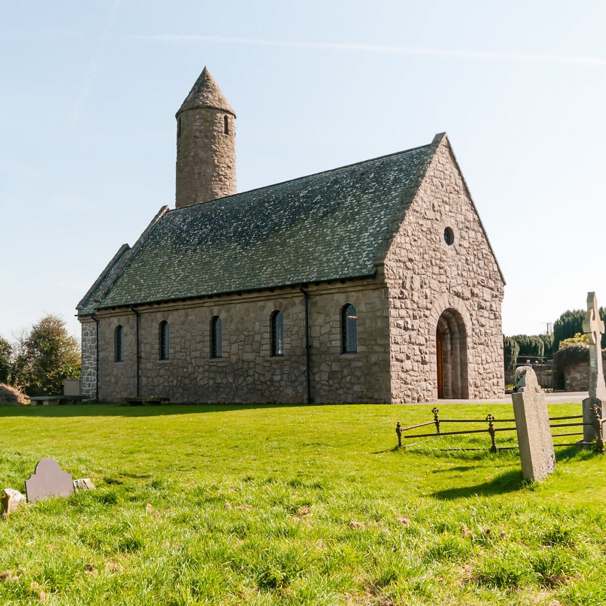 Stone-built Saul Church with round tower and gravestones in the foreground under clear blue sky.