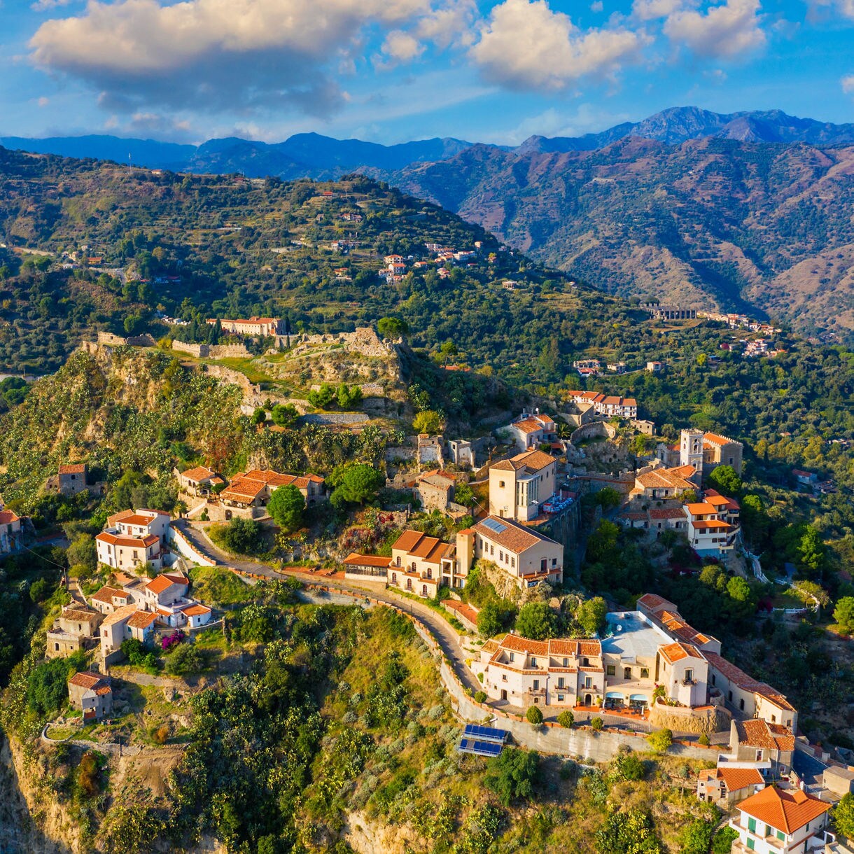 Aerial view of the hilltop village of Savoca in Sicily with clustered stone houses, terracotta roofs, winding roads and surrounding green mountains under a bright sky.