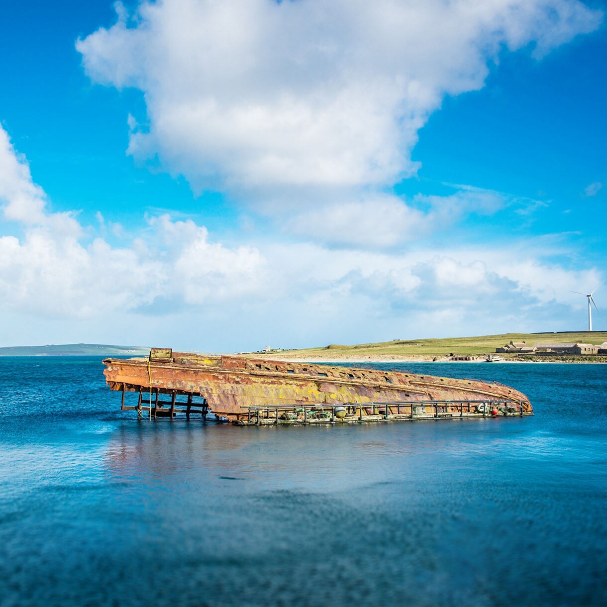Rusted hull of a sunken warship in Scapa Flow, Orkney, partially submerged in calm blue waters with grassy shores and a wind turbine in the distance.