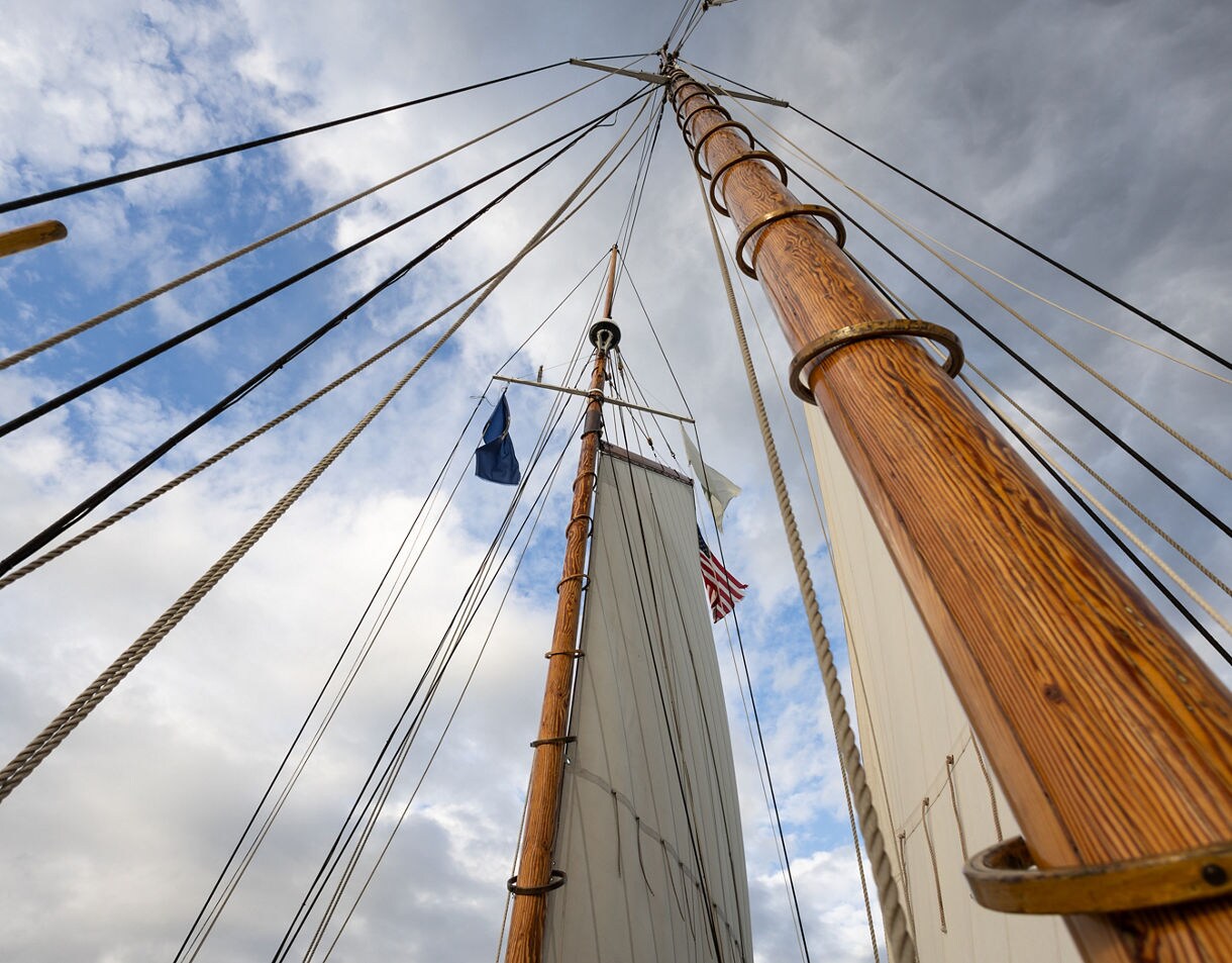 Upward view of tall wooden masts and rigging on a schooner set against a partly cloudy sky.