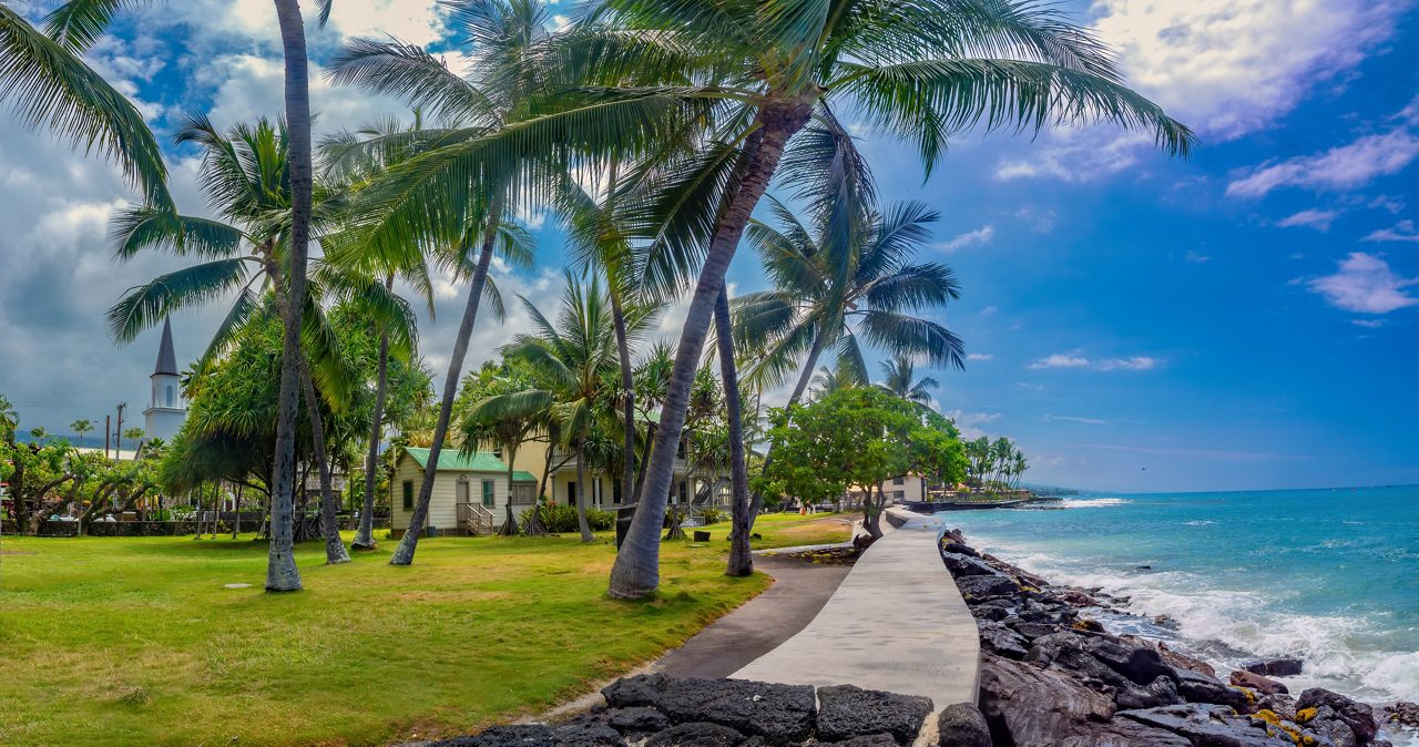 Coastal walkway lined with tall palm trees and ocean waves crashing along black lava rocks in Kailua-Kona, Hawaii.