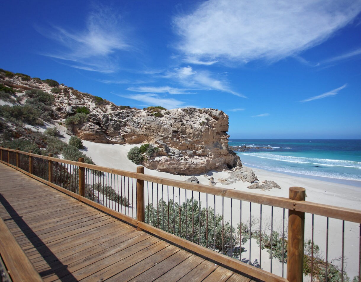 Wooden boardwalk overlooking a sandy beach with rocky cliffs, white dunes and clear blue ocean under bright skies.
