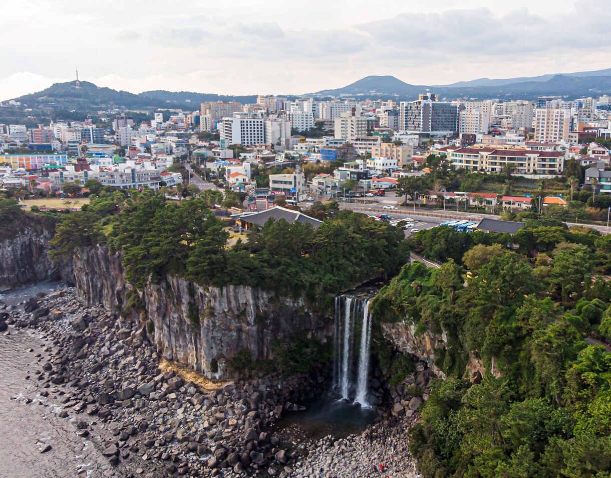 Aerial view of Seogwipo City on Jeju Island, South Korea, showing a tall coastal waterfall cascading from green cliffs into the rocky shoreline below, with city buildings and distant hills in the background.