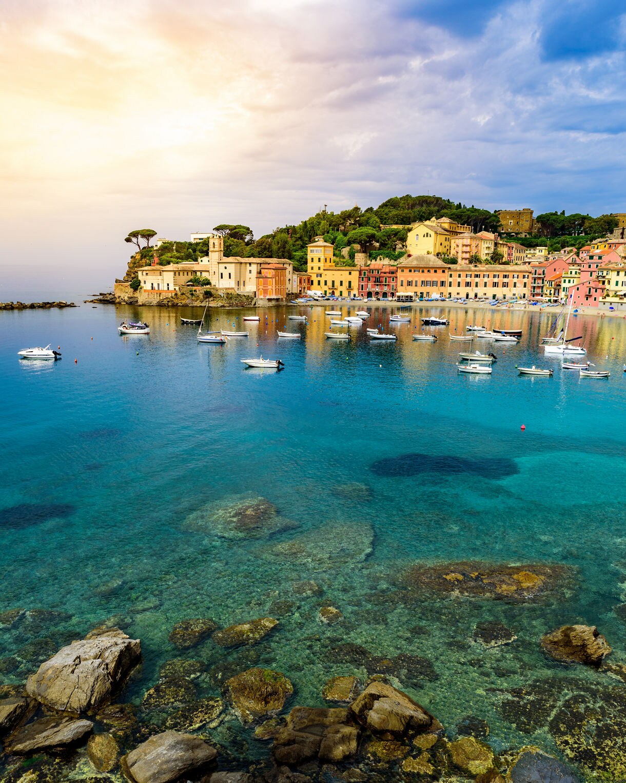 Sunset view of Sestri Levante with colorful waterfront buildings, small boats floating on turquoise water and rocky shallows in the foreground.
