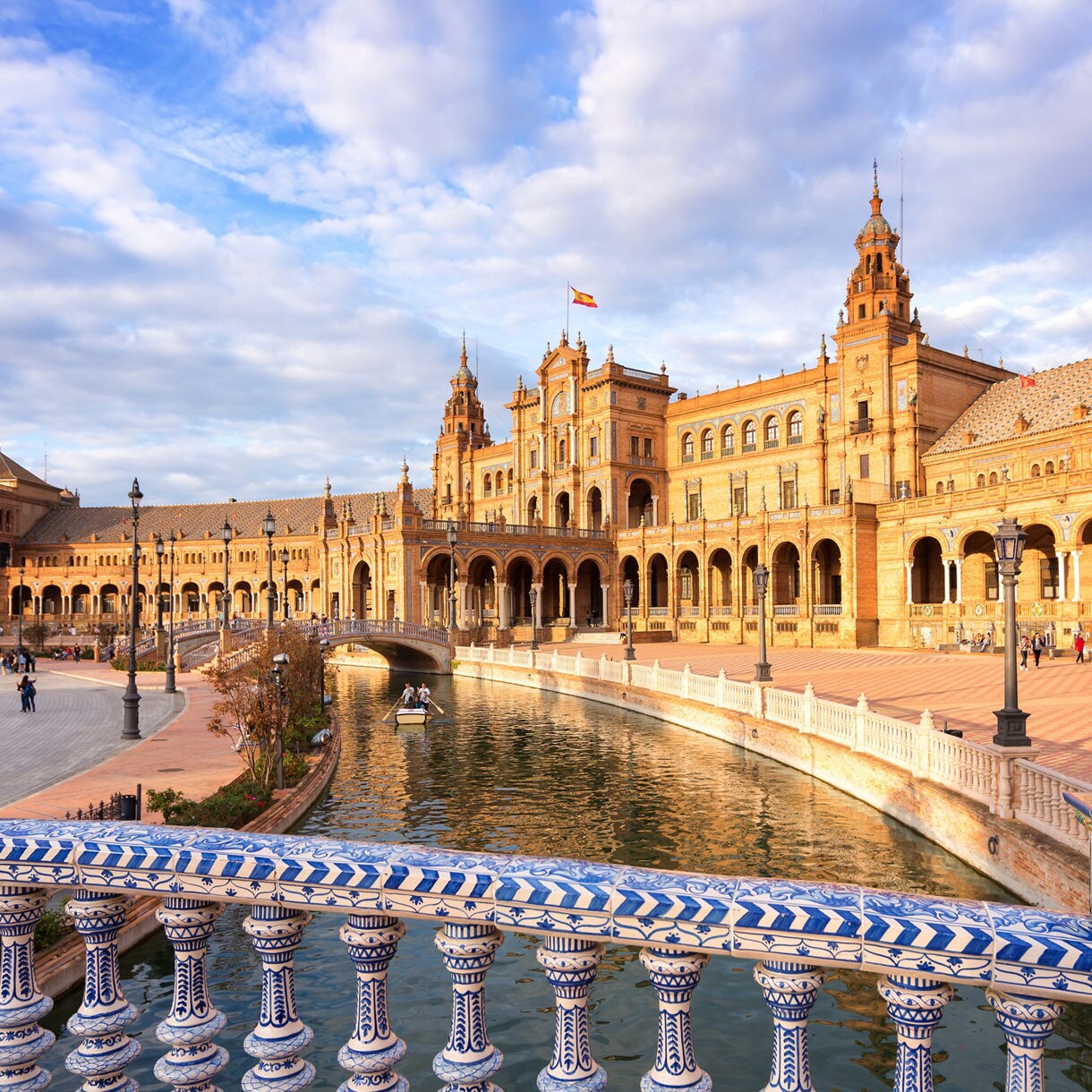 Plaza de España in Seville, Spain, showing its grand curved palace façade, canal with small boats and ornate blue-and-white ceramic balustrade under a partly cloudy sky.