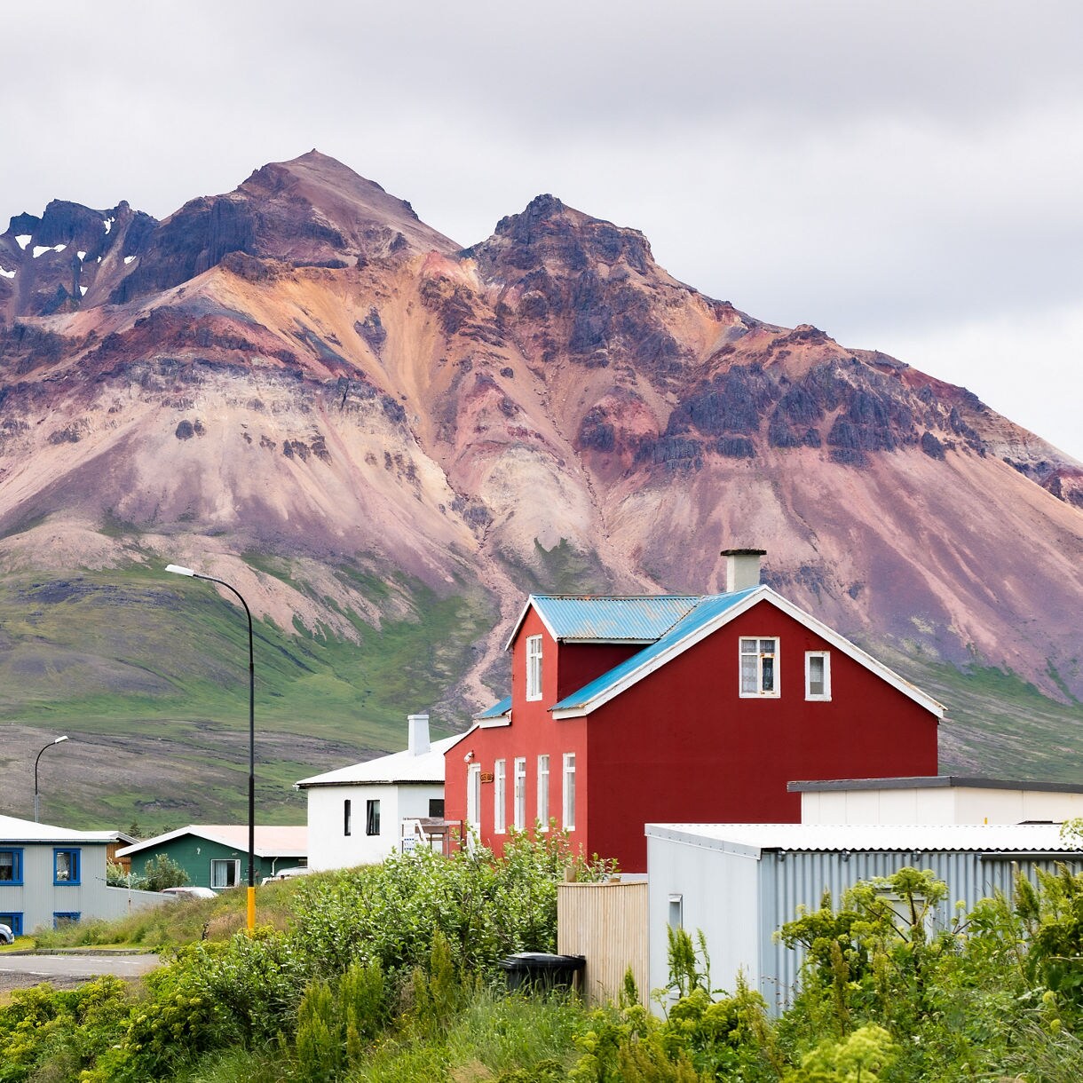A small coastal village featuring a bright red building sits at the base of massive, reddish-brown mountain slopes under an overcast sky.