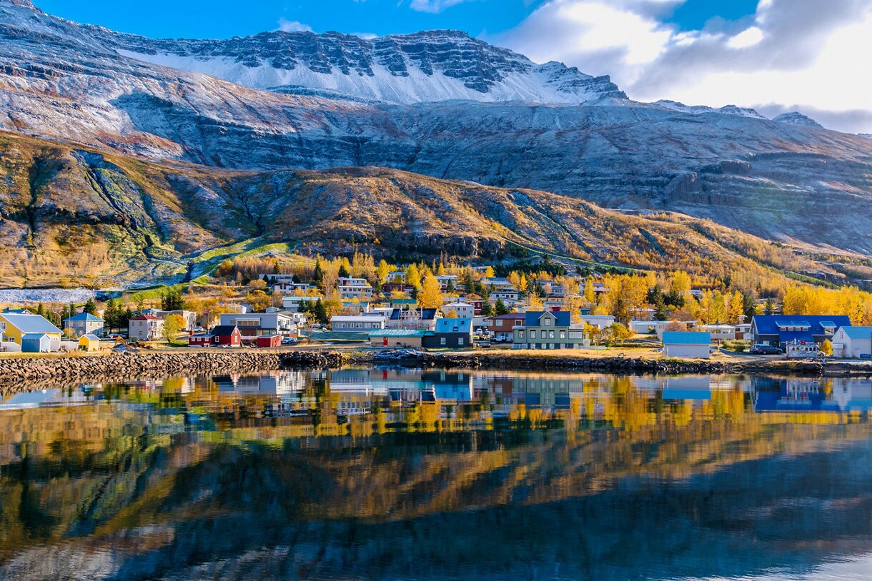 A scenic waterfront view shows colorful wooden buildings of a coastal village reflecting clearly in a calm fjord at the foot of massive, snow-capped mountains.