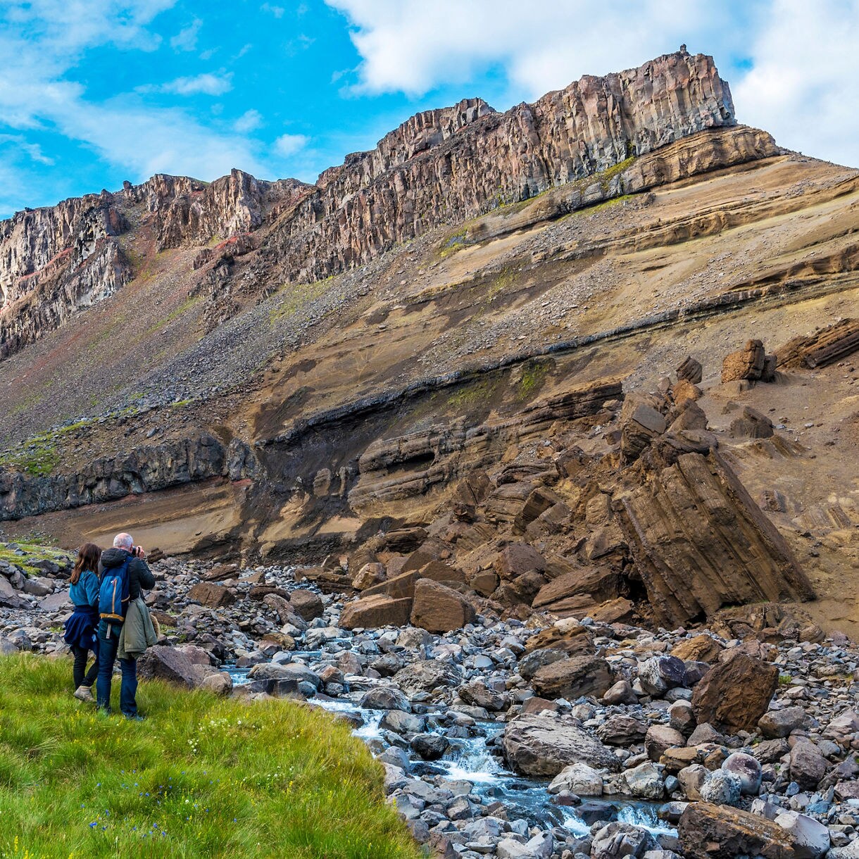 Two hikers stand in a grassy valley beside a rocky stream, taking photos of the tall Hengifoss waterfall cascading down red-striped basalt cliffs under a partly cloudy sky.