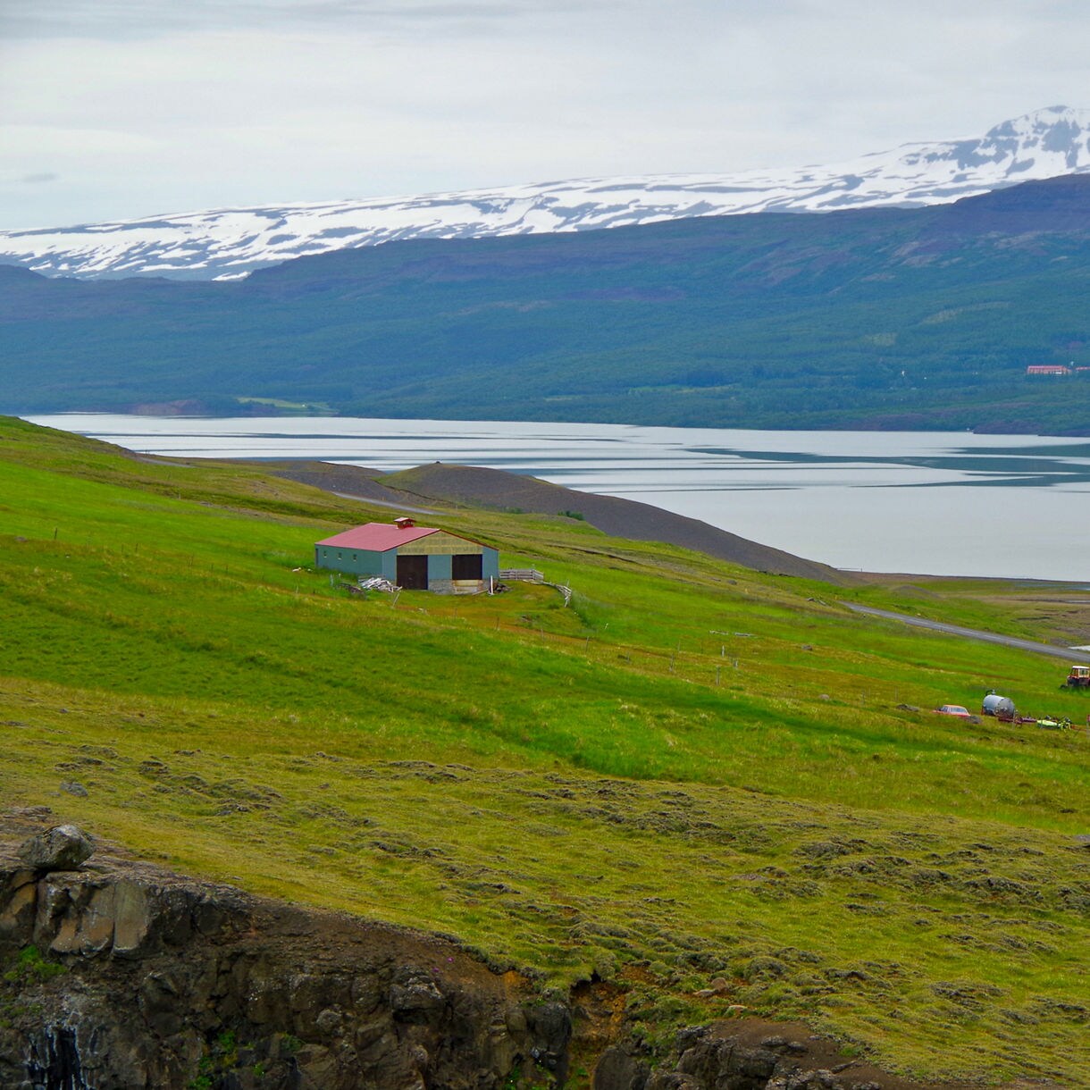 A scenic view overlooks green rolling hills and small farm buildings along the shore of a large, calm lake backed by snow-dusted mountains under an overcast sky.