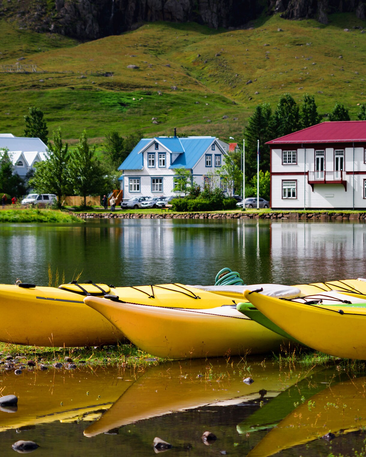 Bright yellow kayaks rest along the grassy shore of a serene lagoon, reflecting the surrounding colorful wooden buildings and a light-blue church set against green mountain slopes under a soft sky.