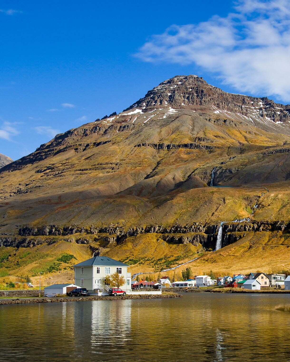 A scenic view shows small, light-colored houses along the edge of a calm fjord beneath a massive, snow-dusted mountain with a cascading waterfall in the background under a bright blue sky.