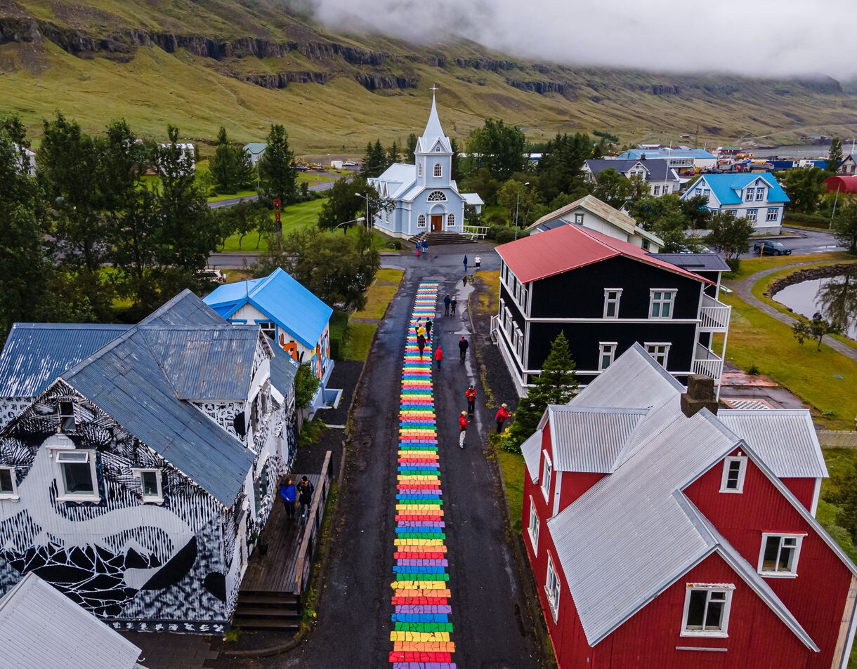 An aerial view shows pedestrians strolling along a street paved with a brightly painted rainbow brick walkway leading toward a picturesque light-blue church framed by lush green mountains.