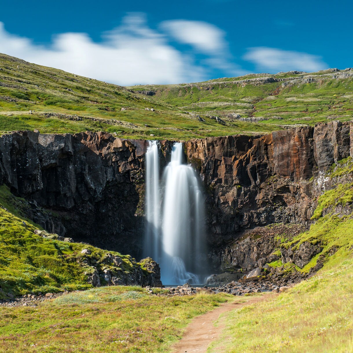 A long-exposure shot captures a silky waterfall cascading down dark basalt rock into a green, sunlit valley beneath a vibrant blue sky with wispy clouds.