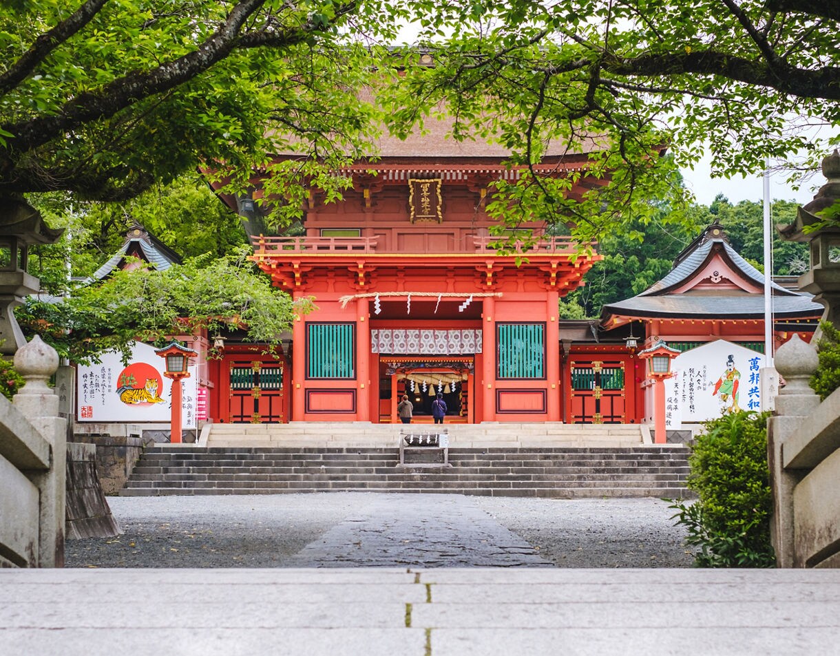 Bright red Shinto shrine with teal lattice windows and ornate details, framed by stone lanterns and overhanging trees, with visitors walking through the main entrance toward the inner courtyard.