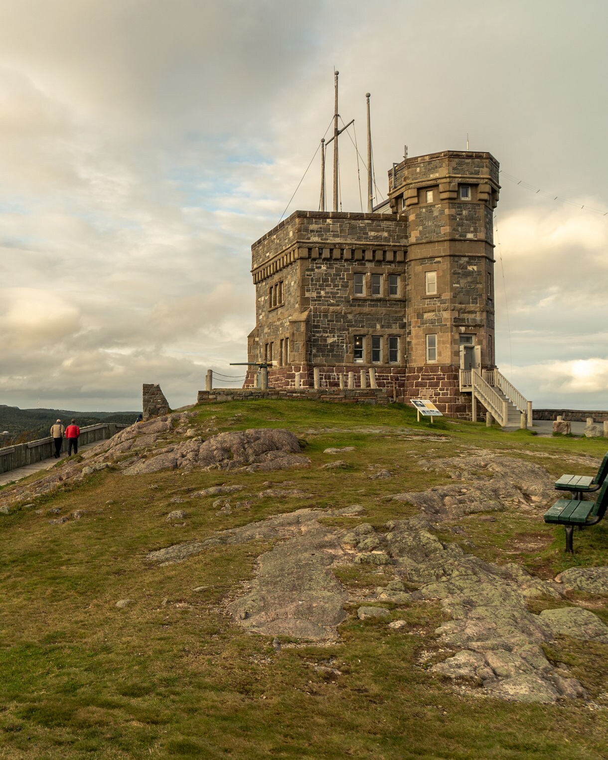 A stone fortress on Signal Hill in St. John’s, Newfoundland, sitting on a grassy rocky slope with two people walking along a wooden boardwalk under a cloudy sky.