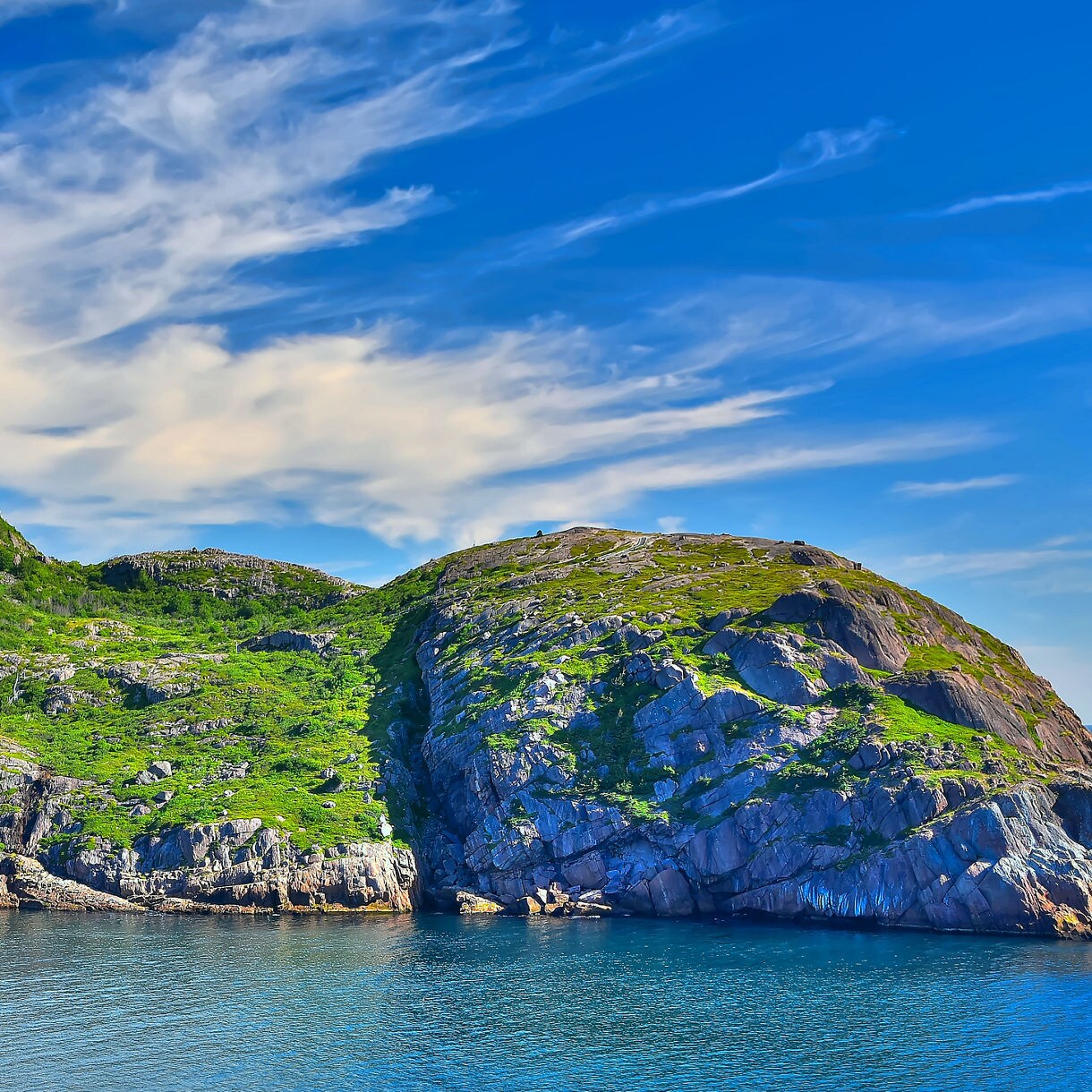 A rugged coastal headland near Signal Hill in Newfoundland, covered in green vegetation with steep rocky faces meeting calm blue ocean water under a vivid sky with wispy clouds.