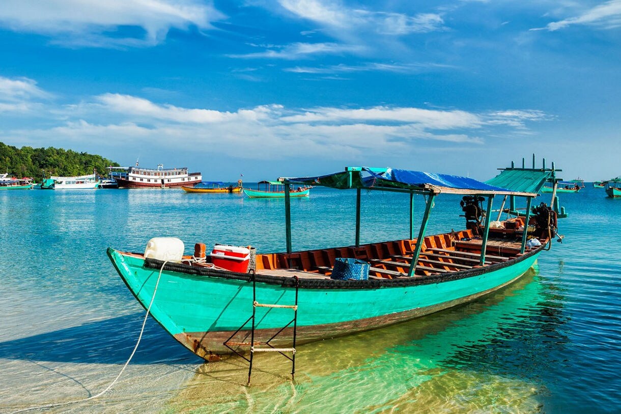 A turquoise wooden boat rests in clear shallow water with more boats and a tree-lined shore under a bright blue sky.