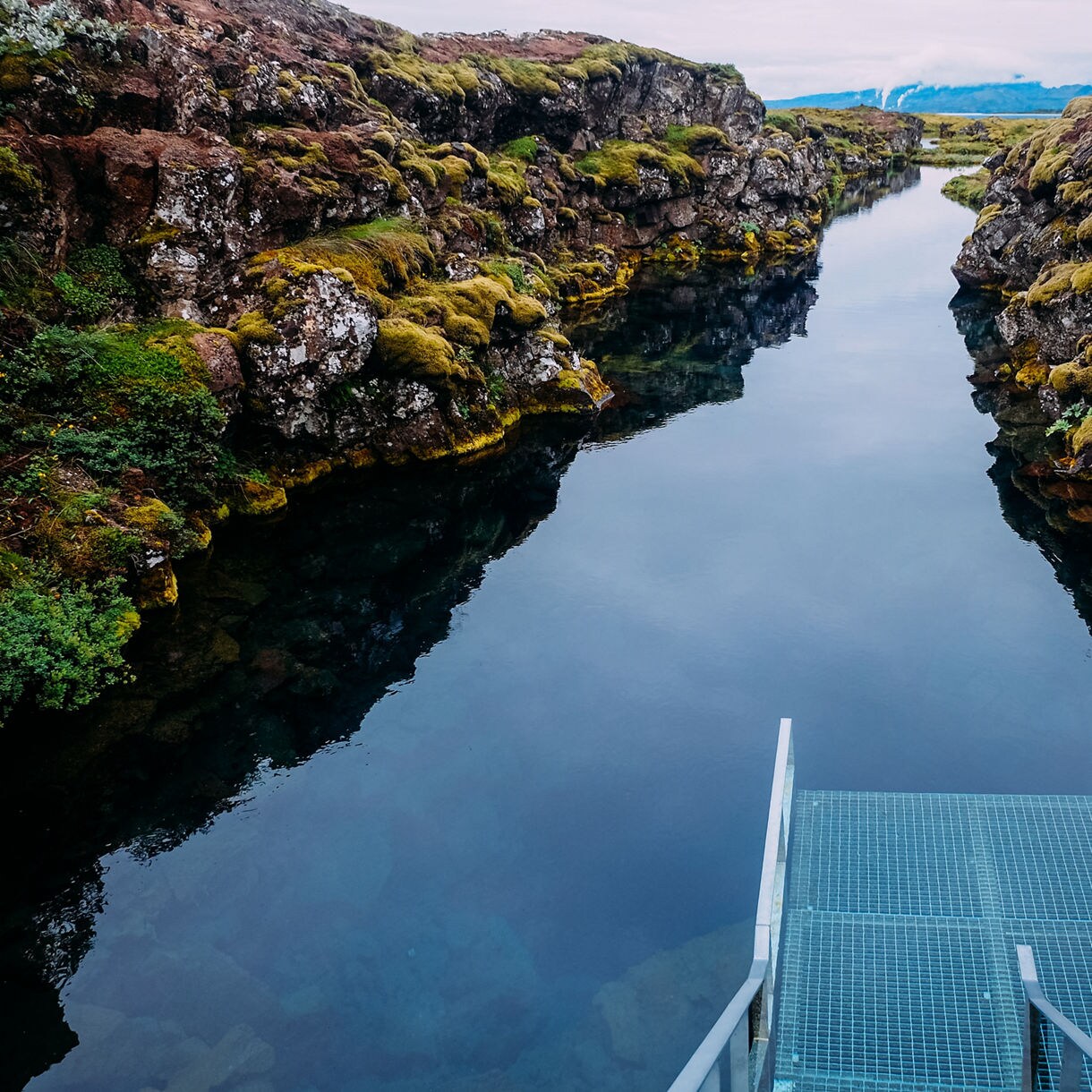 Silfra Fissure in Iceland, a narrow water channel with moss-covered lava rock walls and a metal diving platform leading into the transparent blue water.